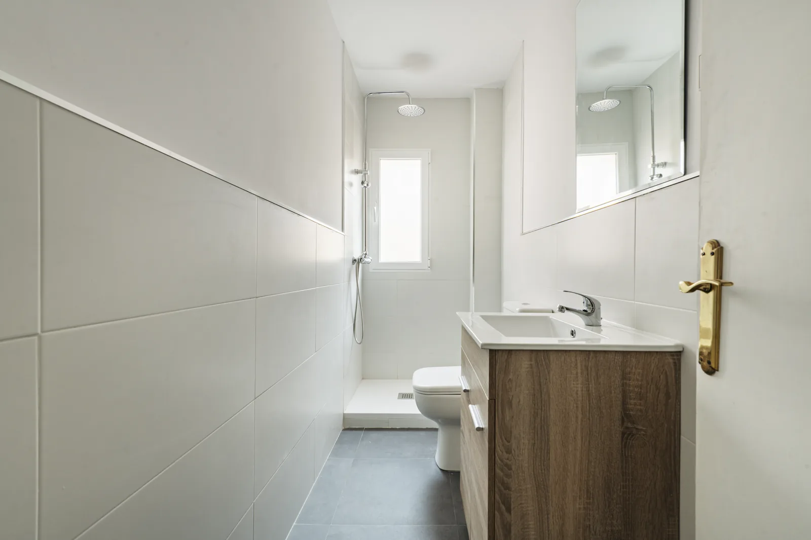 Casa de banho pequena com duche walk-in ao fundo, azulejo branco a meia altura e móvel de lavatório em madeira